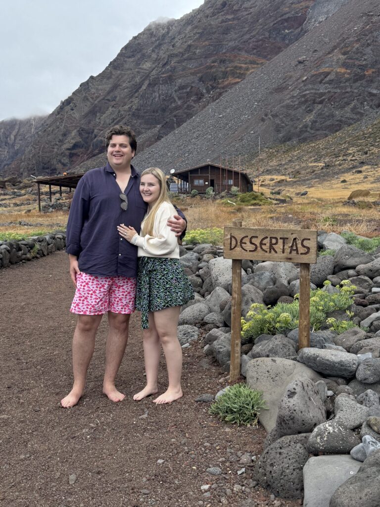 A couple barefoot on a stony path embraces in front of a wooden hut and "DESERTAS" sign, with barren volcanic mountains rising under cloudy skies on Desertas Islands, an experience by Luxury Charter Madeira.