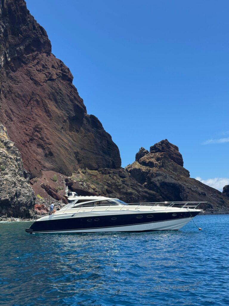 A sleek white and navy yacht floats on clear blue water at the base of towering, rugged volcanic cliffs under a bright, cloudless sky, an experience in a Luxury Yacht Charter near Desertas Island