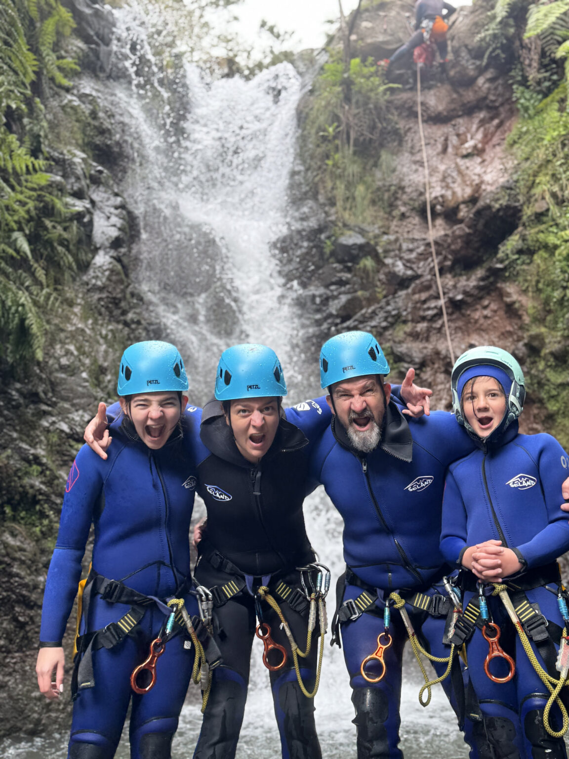 A joyful family of four canyoneers poses arm-in-arm in stream waters with an instructor rappelling a waterfall above in Porto Novo, Camacha, during a Beginner Level 1 tour with Go Explore Madeira.