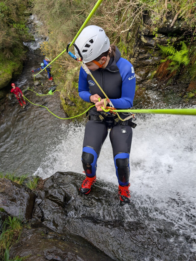 A female canyoneer prepares her ropes at the edge of a small waterfall in Porto Novo, Camacha, with group members visible below during a Beginner Level 1 tour with Go Explore Madeira.