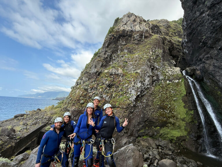 A group of five canyoneers in blue wetsuits and helmets pose triumphantly with peace signs and thumbs up beside a thin waterfall cascading from mossy volcanic cliffs into the Atlantic Ocean, with distant Desertas Islands and clouds on the horizon in Madeira during an Advanced Level 3 canyoning experience with Go Explore Madeira.
