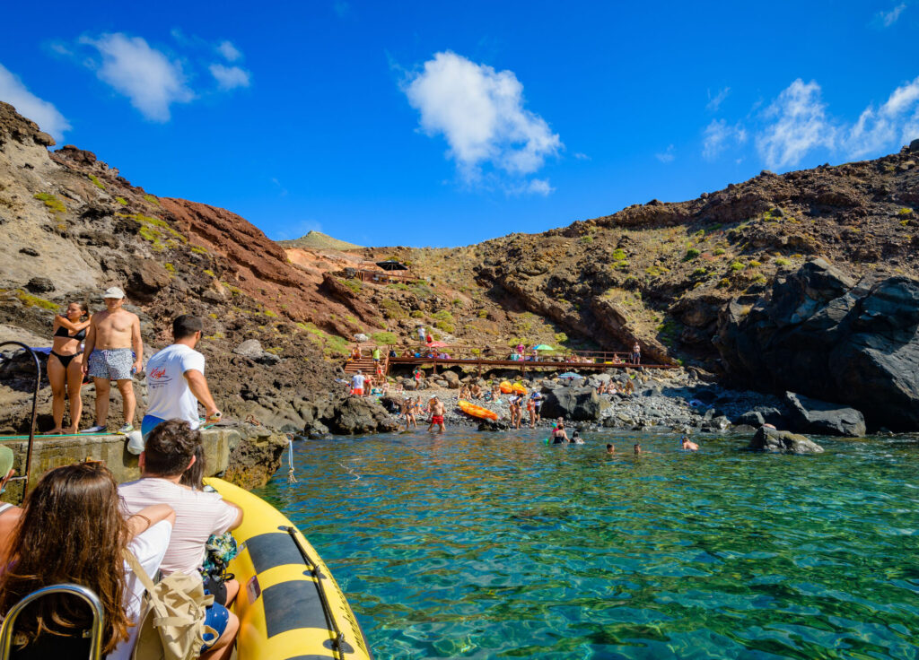 PR8 Madeira: The Vereda da Ponta de São Lourenço Walking Guide 5 Madeira Sea Emotions boat tour filled with people on a dock Cais do Sardinha bay where there is people sunbathing at the rocky beach and people swimming in the water.