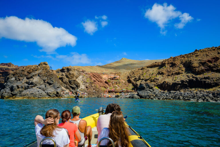 The sea, rocky land and blue cloudy sky on the horizon, and People on a Madeira Sea Emotions boat tour arriving at the Cais do Sardinha.