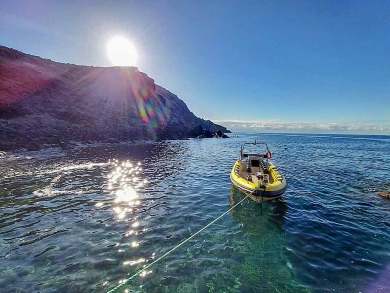 A yellow Madeira Sea Emotions RIB boat rests on crystal-clear water at Ponta de São Lourenço, with the sun setting behind rugged cliffs and casting rainbow flares across the calm cove.