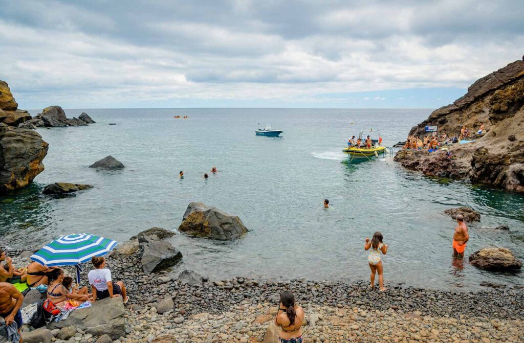 Crowds relax on the rocky shore and swim in the calm cove at Cais do Sardinha, while a yellow Madeira Sea Emotions tour boat is taking more people to do some touring on the coast.