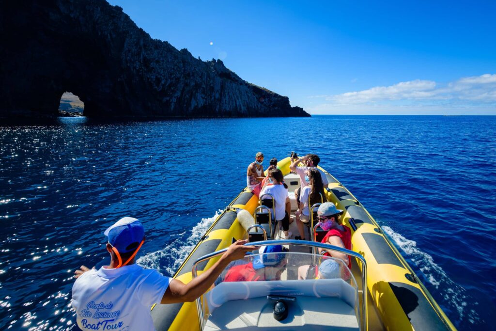 A yellow RIB boat full of tourists and a guide in a cap cruises past Ponta de São Lourenço's towering dark cliffs and a rock arch under bright blue skies, a boat tour experience by Madeira Sea Emotions.
