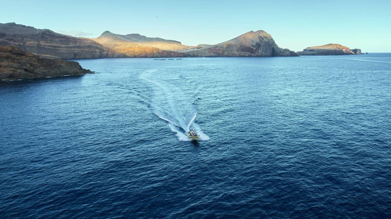 A boat leaves a white wake across blue waters off Ponta de São Lourenço's dramatic, sunlit volcanic cliffs and headlands, a boat tour experience by Madeira Sea Emotions.