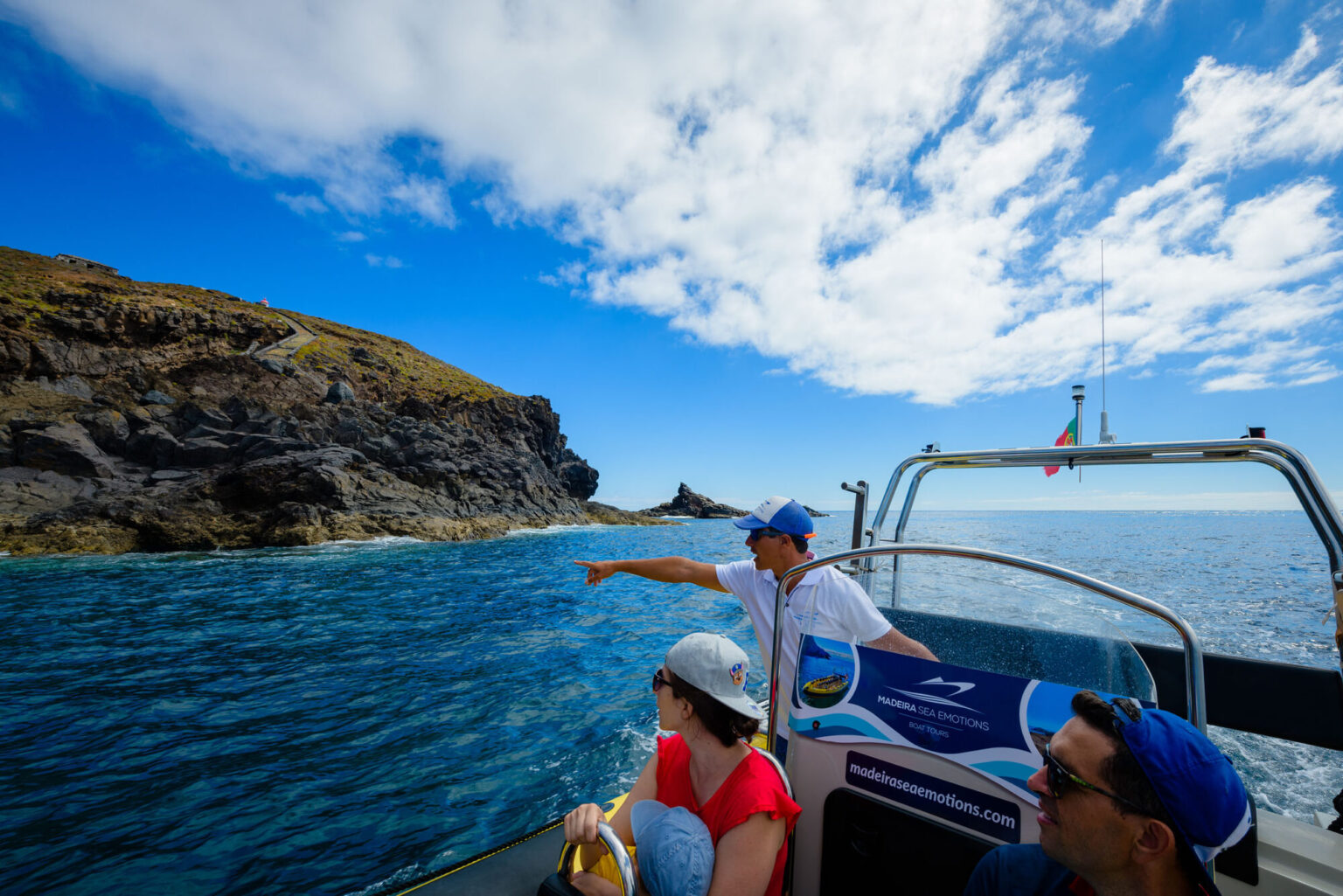 A guide in a cap points from a blue Madeira Sea Emotions RIB boat toward a path on volcanic cliffs under partly cloudy skies, a boat tour experience by Madeira Sea Emotions.
