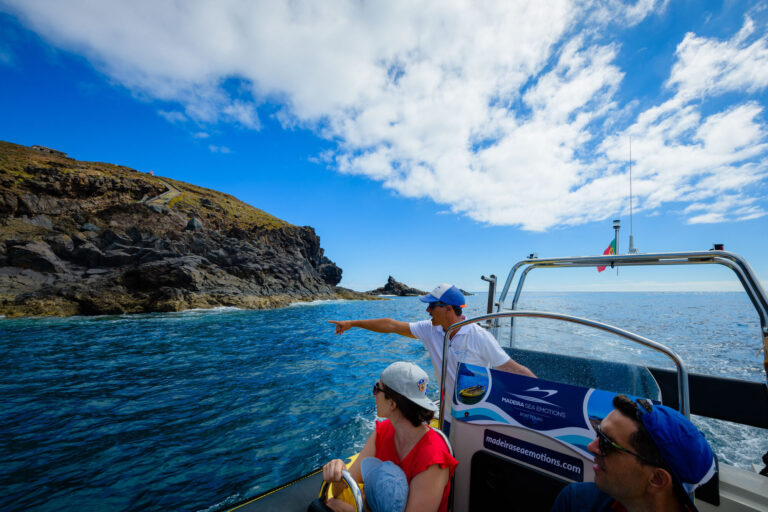 A guide in a cap points from a blue Madeira Sea Emotions RIB boat toward a path on volcanic cliffs under partly cloudy skies, a boat tour experience by Madeira Sea Emotions.