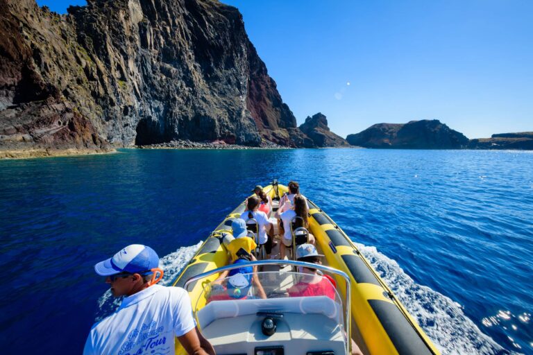 A yellow RIB boat full of tourists and a guide in a cap cruises past Ponta de São Lourenço's towering dark cliffs and a rock arch under bright blue skies, a boat tour experience by Madeira Sea Emotions.