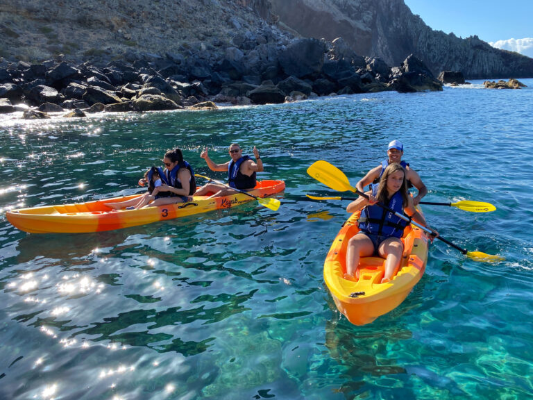 Two orange kayaks paddle along Ponta de São Lourenço's dark volcanic coastline, with life-vested explorers enjoying the turquoise waters and sunny skies, a Kayaking experience by Madeira Sea Emotions.