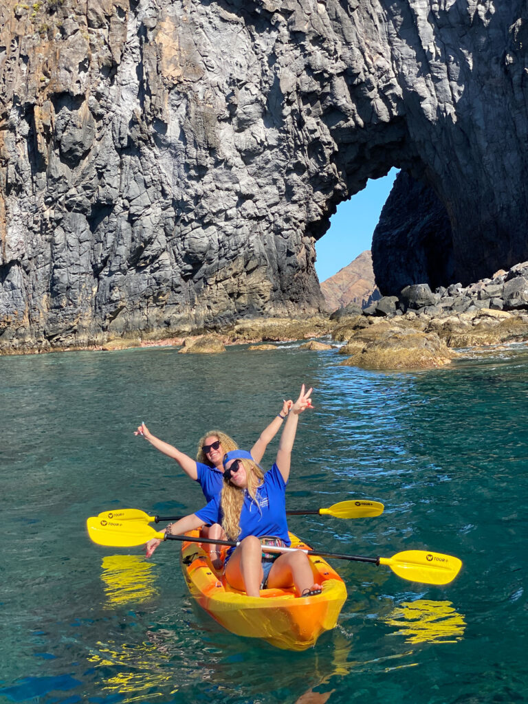 2 girls Kayaking in a full day pack of fun along Ponta de São Lourenço's rugged volcanic coast, framed by a massive rock archway in a kayaking Madeira Sea Emotions experience.