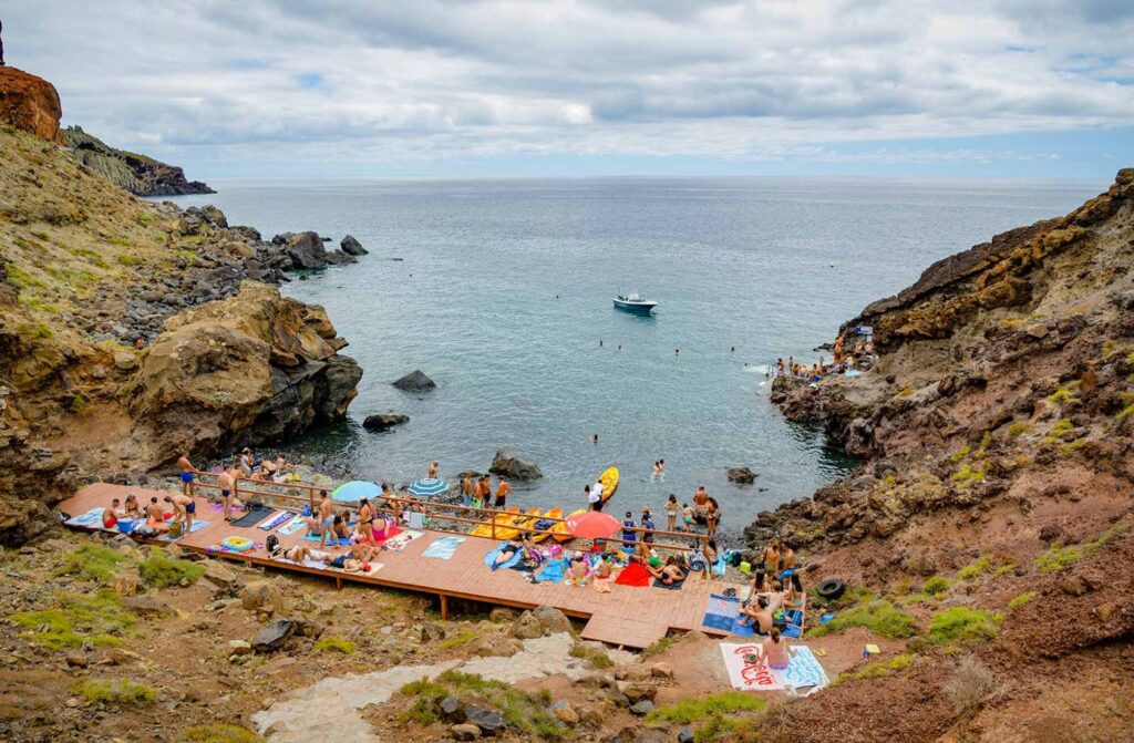 PR8 Madeira: The Vereda da Ponta de São Lourenço Walking Guide 3 Cais do Sardinha with people relaxing taking sunbathing and swimming on the sea and a yellow tour boat of Madeira Sea Emotions leaving with people for another tour around the Madeira Coast.