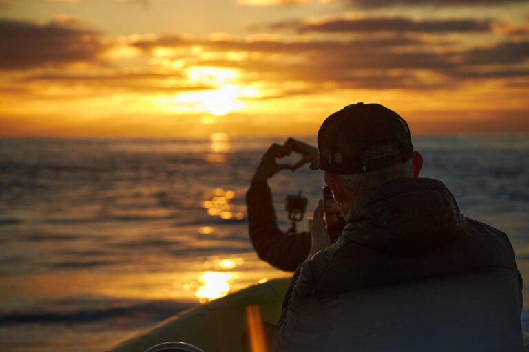two people on a boat one doing a shape heart with hands and another filming it with a mobile phone with a sunset over the sea in the back during a boat tour experience by Madeira Sea Emotions.
