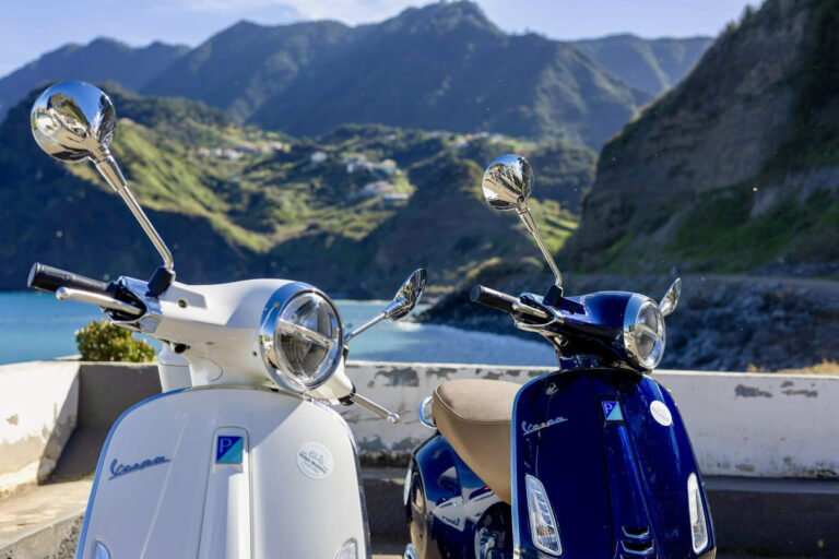 A close up of Two Vespa scooters,a blue one and a white one by Vespa Madeira Scooter Rent parked and on the back the Sea and mountains of Madeira Island. These are the type of scooters offered for rent to explore the Madeira island’s winding mountain roads and scenic viewpoints by Vespa Madeira Scooter Rent.