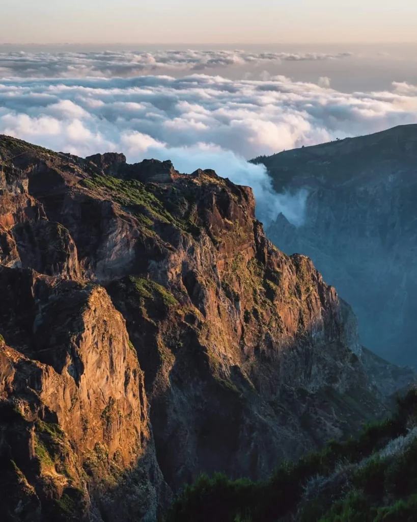East vs. West Madeira: Which 4x4 Jeep Tour is Best? 7 Dramatic sunrise view of rugged cliffs in eastern Madeira, with golden light on the rocky slopes and a thick sea of clouds filling the deep valley below. A Jeep tour experience by Sea La Vie Mountain Expedition.