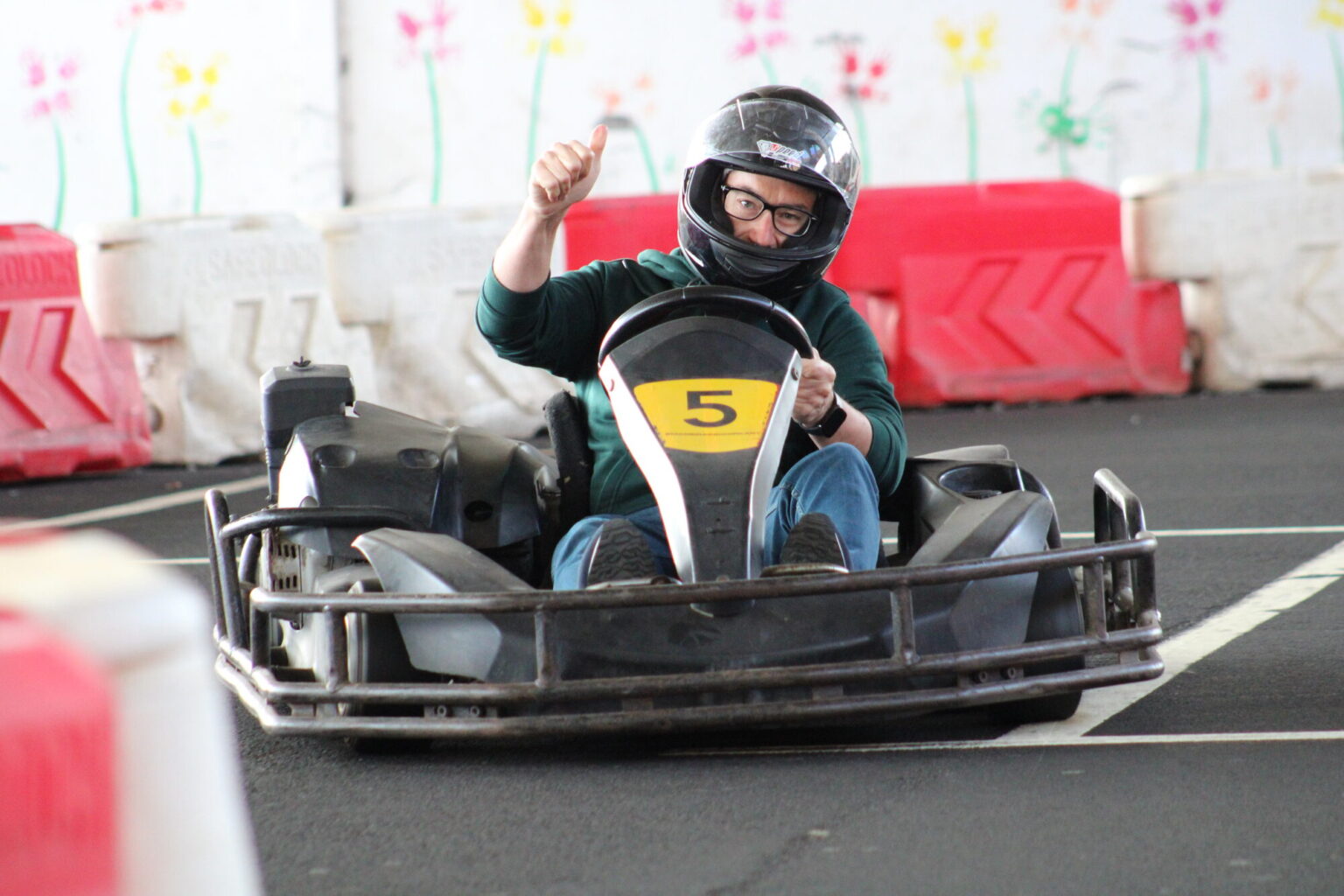 Indoor go‑kart driver in kart number 5 giving a thumbs-up while racing on a track located under the Madeira Airport with safety barriers and colorful wall decorations in the background.An experience by Go Kart Madeira