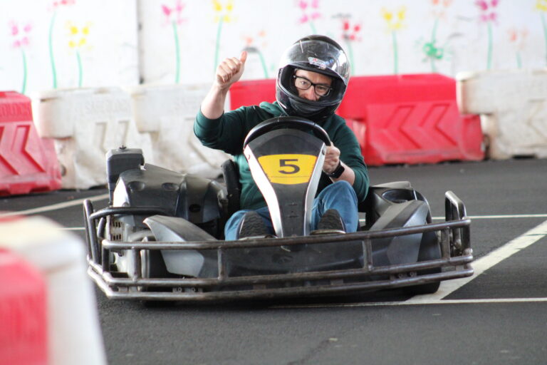 Indoor go‑kart driver in kart number 5 giving a thumbs-up while racing on a track located under the Madeira Airport with safety barriers and colorful wall decorations in the background.An experience by Go Kart Madeira