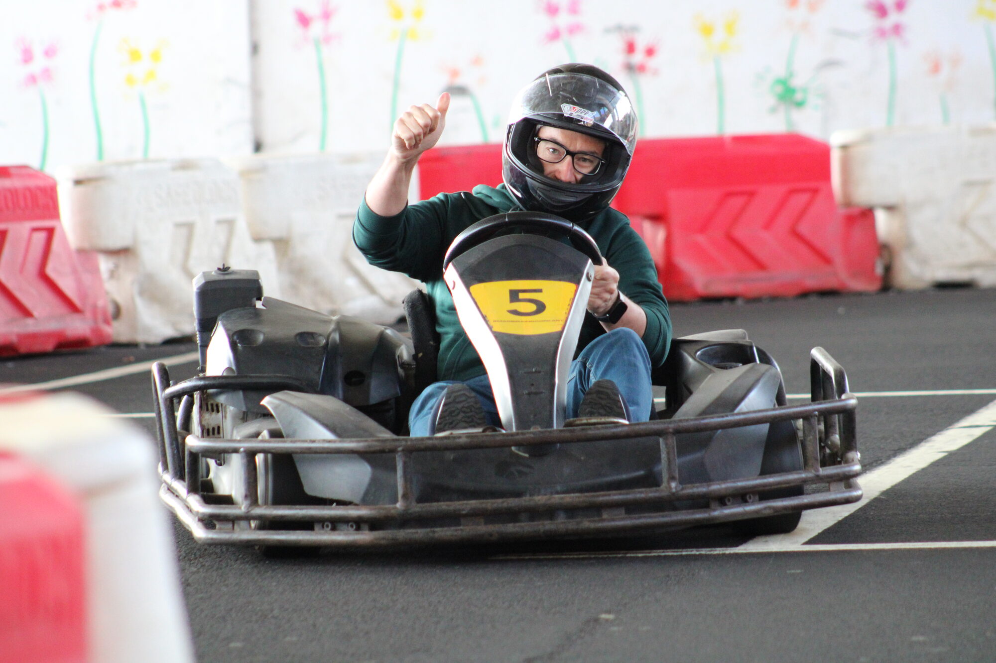 Indoor go‑kart driver in kart number 5 giving a thumbs-up while racing on a track located under the Madeira Airport with safety barriers and colorful wall decorations in the background.An experience by Go Kart Madeira