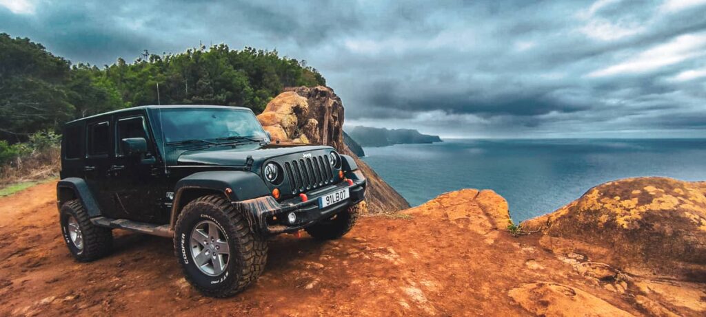 Black Jeep Wrangler parked on a cliff-edge dirt track in Madeira, with rugged coastal cliffs, forested slopes and a dramatic cloudy sky over the Atlantic Ocean., during an offroad tour by Sea La Vie Mountain Expedition.