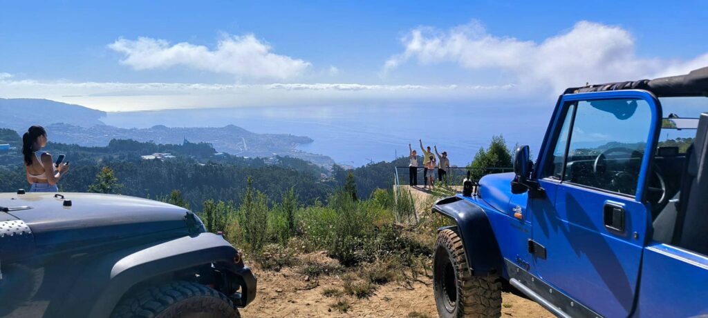East vs. West Madeira: Which 4x4 Jeep Tour is Best? 1 Panoramic hilltop viewpoint in Madeira with two Jeeps parked in the foreground, a woman filming on the left, and a group raising their arms on a distant platform overlooking the coastline and Atlantic Ocean., during an offroad tour by Sea La Vie Mountain Expedition.