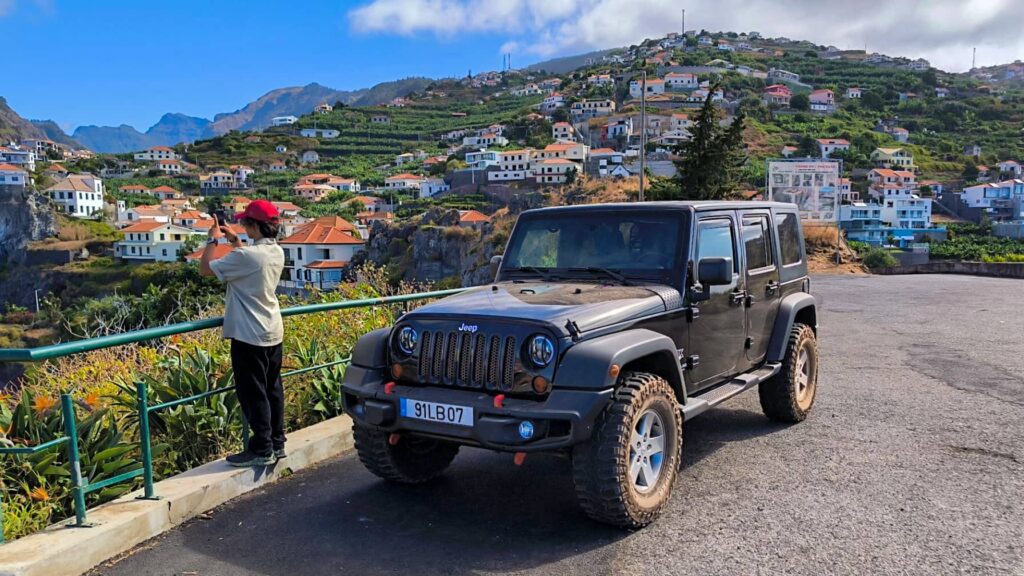 East vs. West Madeira: Which 4x4 Jeep Tour is Best? 9 Black Jeep Wrangler parked at a roadside viewpoint in Madeira, with a person taking photos over a green railing and a hillside village of terraced houses and mountains in the background. A Jeep tour experience by Sea La Vie Mountain Expedition.