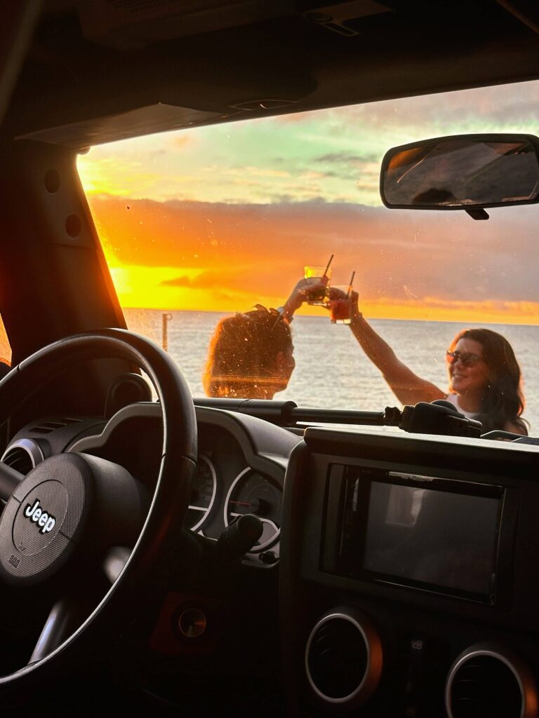 Interior view from a Jeep looking out toward the ocean at sunset, with two people outside clinking drinks against a vibrant orange and yellow sky over the water.A Jeep tour experience by Sea La Vie Mountain Expedition.