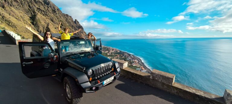 Family enjoying a scenic stop with a black Jeep Wrangler at a cliffside viewpoint in Madeira, doors open and arms raised, overlooking a coastal town and the bright blue Atlantic Ocean. A Jeep tour experience by Sea La Vie Mountain Expedition.