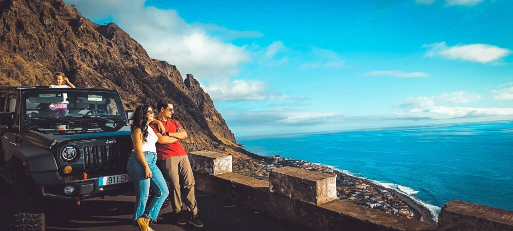East vs. West Madeira: Which 4x4 Jeep Tour is Best? 8 Couple standing by a black Jeep Wrangler at a scenic cliffside viewpoint in Madeira, overlooking a coastal town, steep rocky mountains and the turquoise Atlantic Ocean under a bright blue sky. Madeira North & West Mountain Jeep Expedition Tour Adventure by Sea La Vie Mountain Expedition.