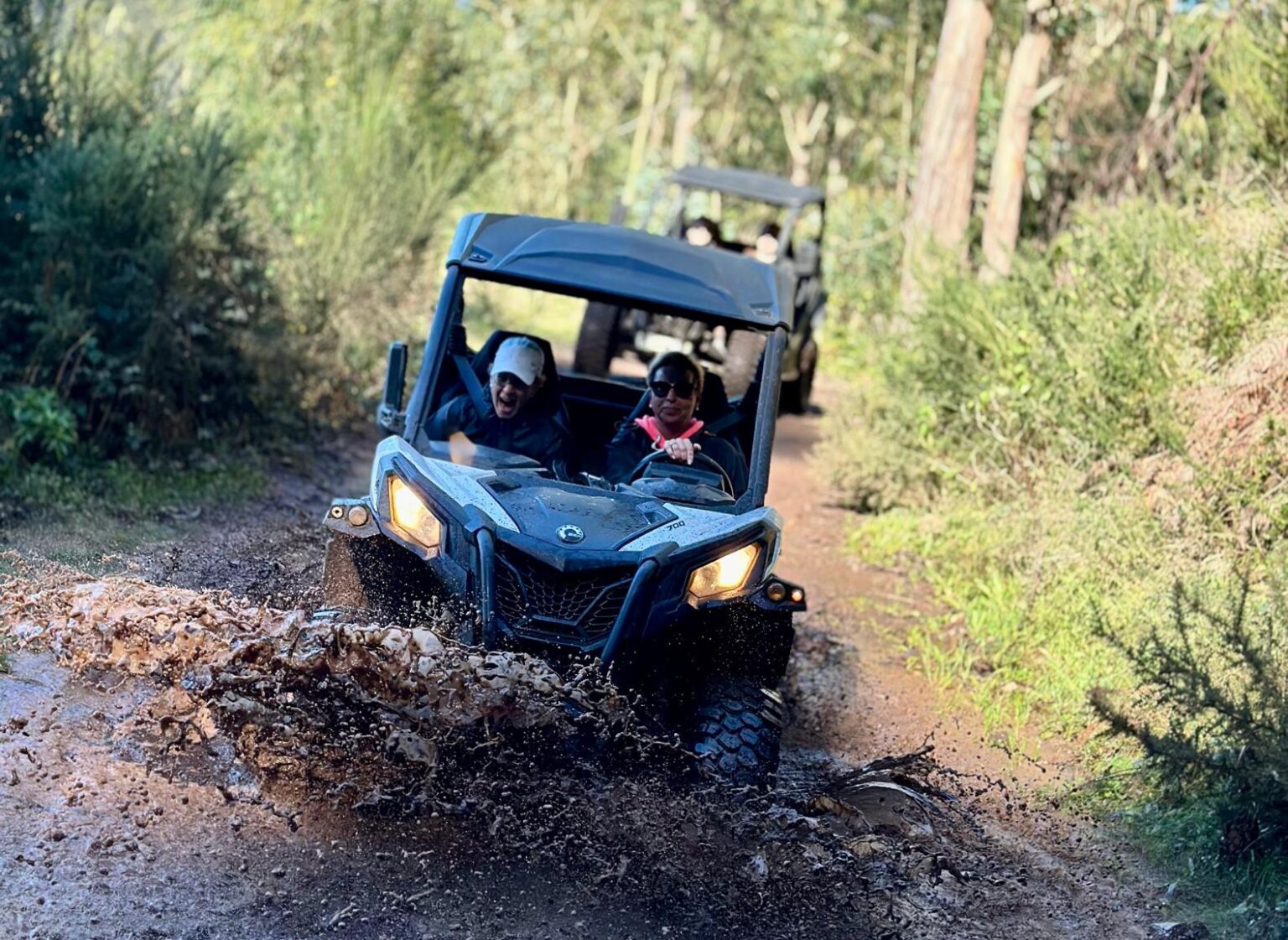 Buggy splashing through a muddy puddle on a forest trail near Câmara de Lobos, with another vehicle following behind among the green trees, during an adventure buggy tour by Madeira Radical.