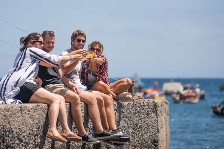 Friends toasting poncha drinks while relaxing by the sea during the Bar hopping and Poncha Workshop, an experience by Wine Tours Madeira.