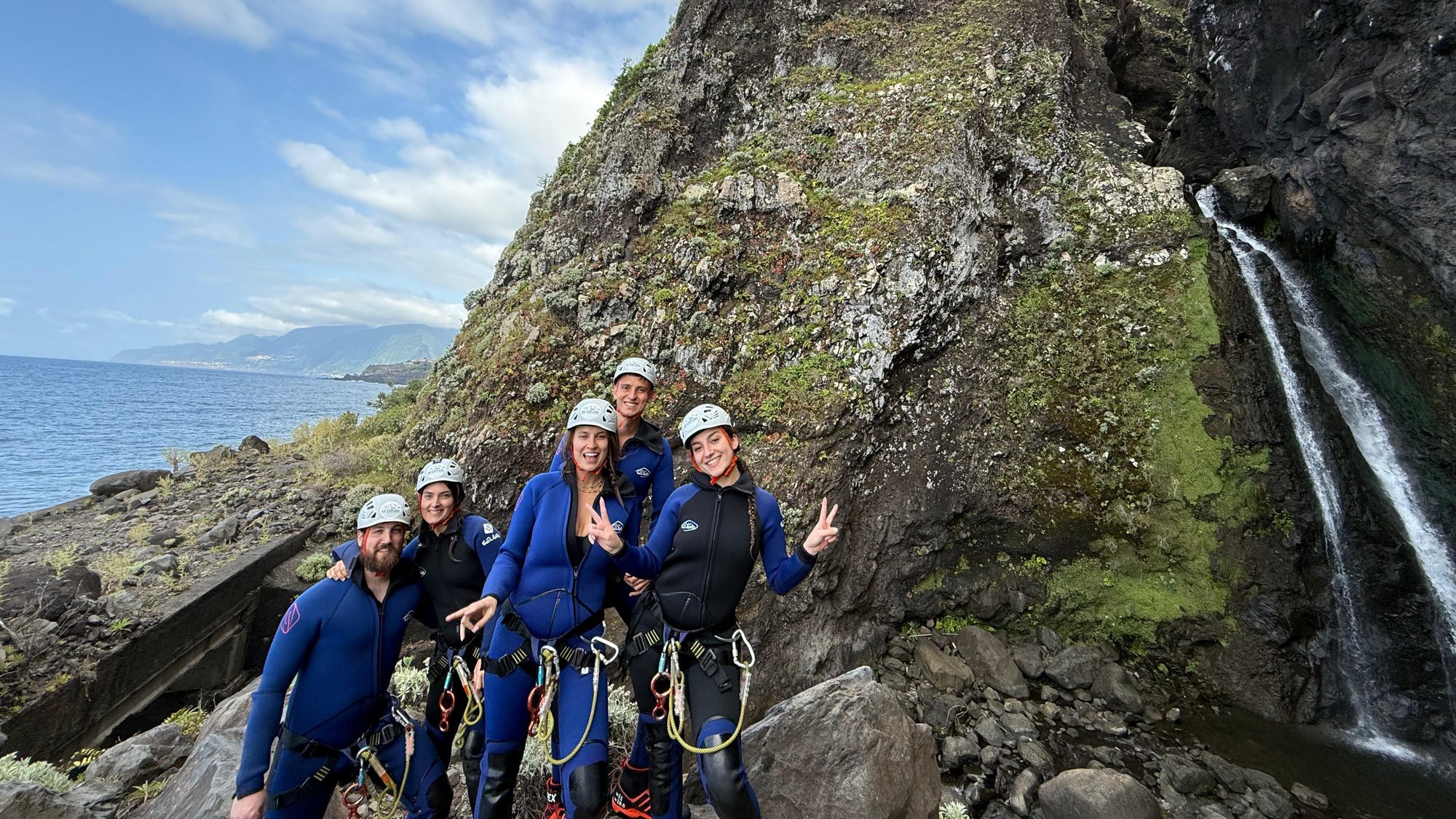 Canyoning in Madeira, A Practical Guide 1 A group of five canyoneers in blue wetsuits and helmets pose triumphantly with peace signs and thumbs up beside a thin waterfall cascading from mossy volcanic cliffs into the Atlantic Ocean, with distant Desertas Islands and clouds on the horizon in Madeira during an Advanced Level 3 canyoning experience with Go Explore Madeira.