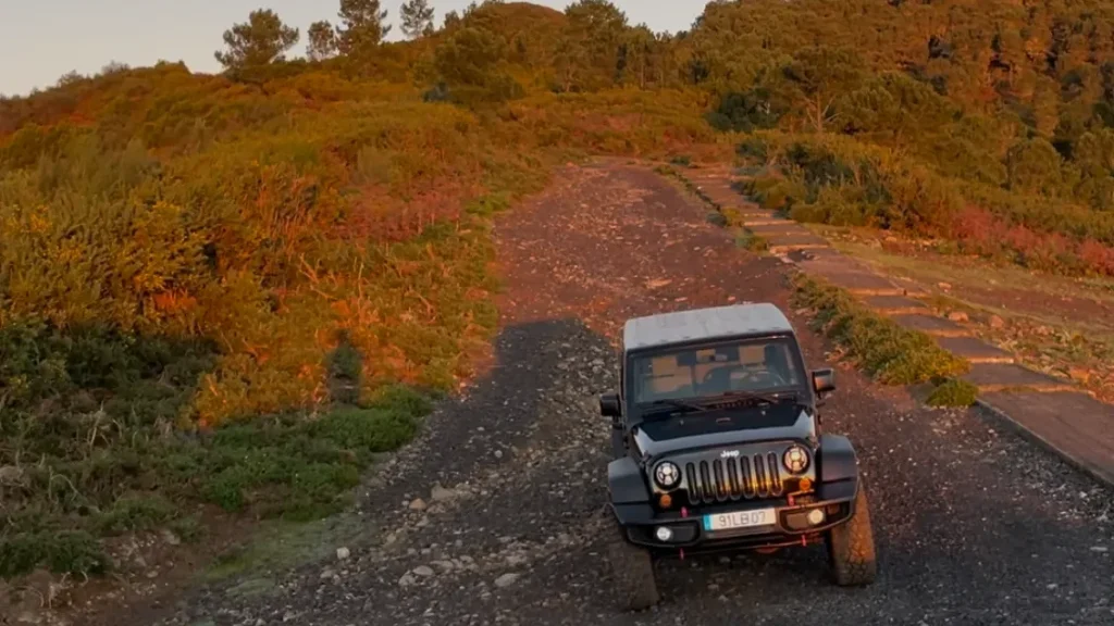 East vs. West Madeira: Which 4x4 Jeep Tour is Best? 3 Sunset off‑road scene with a black Jeep Wrangler parked on a rocky gravel track, surrounded by green and orange-lit hillsides, under a clear sky with the moon visible above.An experience by Sea La Vie Mountain Expedition.