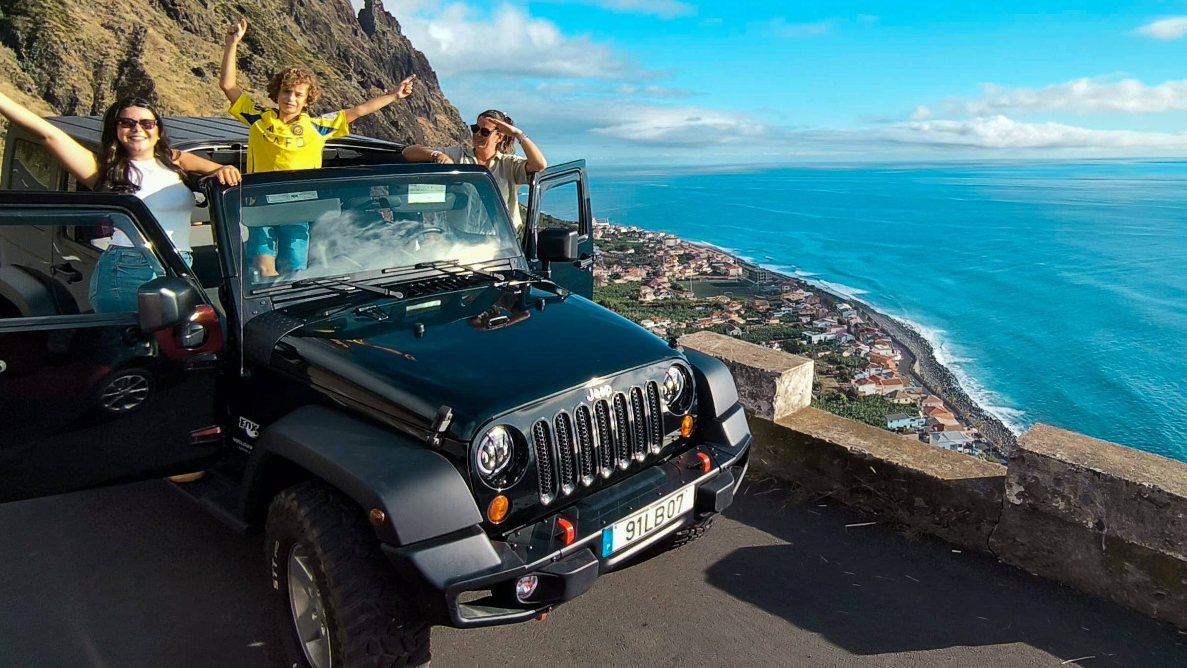 Canyoning in Madeira, A Practical Guide 6 Family enjoying a scenic stop with a black Jeep Wrangler at a cliffside viewpoint in Madeira, doors open and arms raised, overlooking a coastal town and the bright blue Atlantic Ocean. A Jeep tour experience by Sea La Vie Mountain Expedition.