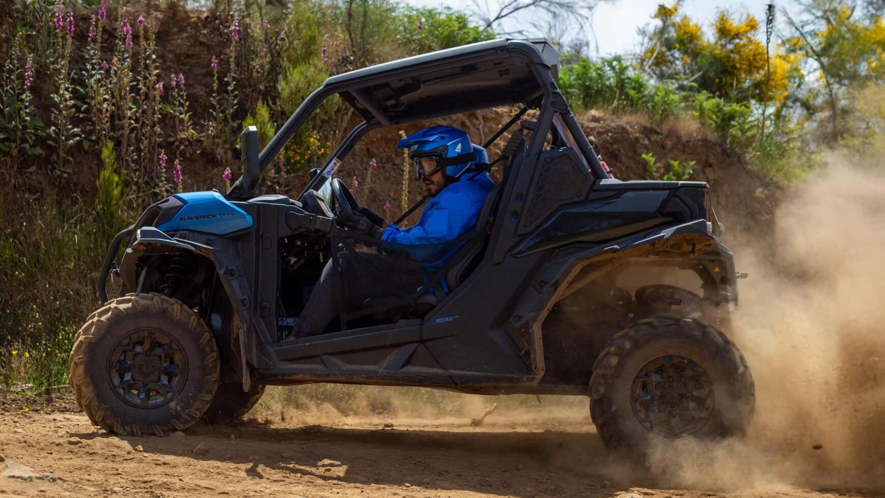 Which Buggy Tour in Madeira Is Right for You 4 A driver in a blue helmet powers a Can-Am Maverick buggy through a dusty corner, kicking up a cloud of dirt on a rugged off-road track lined with wildflowers and greenery in Madeira Laurissilva forest, during a Let's Buggy's Laurissilva experience in Madeira.