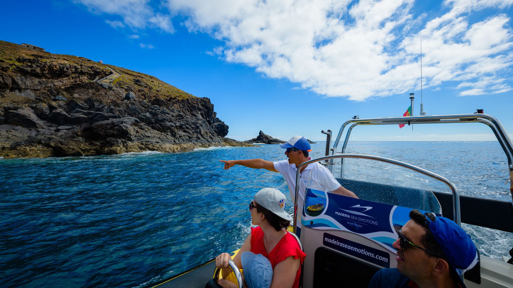 Best boat trips in Madeira 2 A guide in a cap points from a blue Madeira Sea Emotions RIB boat toward a path on volcanic cliffs under partly cloudy skies, a boat tour experience by Madeira Sea Emotions.