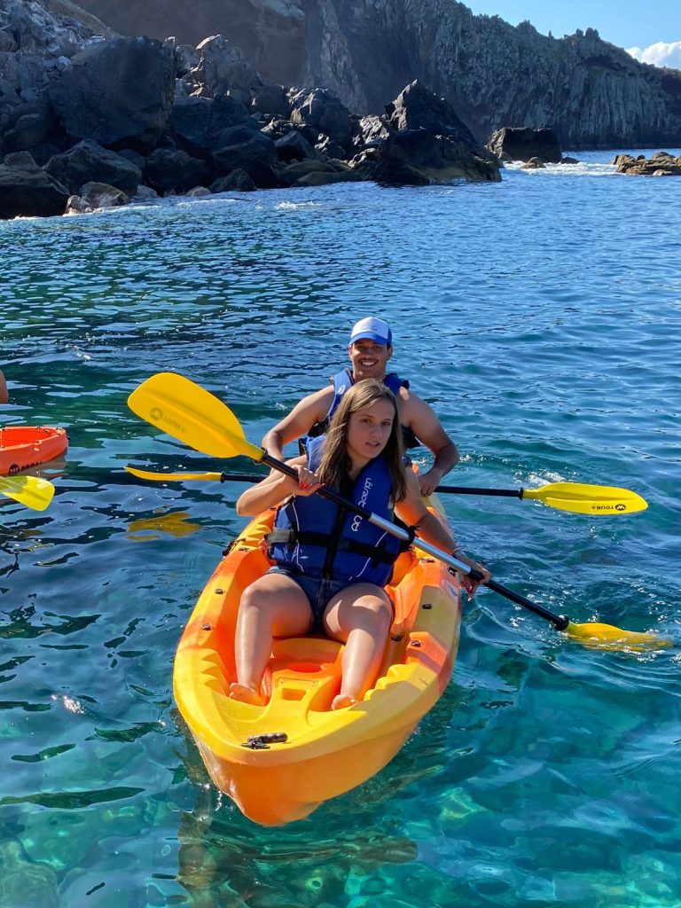 Two orange kayaks paddle along Ponta de São Lourenço's dark volcanic coastline, with life-vested explorers enjoying the turquoise waters and sunny skies, a Kayaking experience by Madeira Sea Emotions.