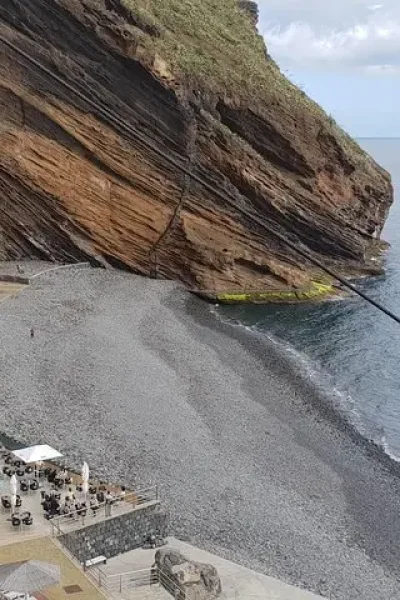 Grey pebble beach and beach bar tucked beneath a layered rock cliff on Madeira's south coast
