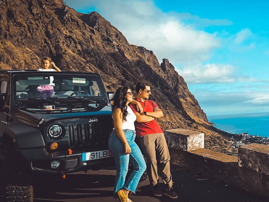 Group posing with a black Jeep Wrangler at a scenic roadside viewpoint in Madeira, with steep rocky cliffs behind them and a coastal town and blue Atlantic Ocean below.An experience by Sea La Vie Mountain Expedition.