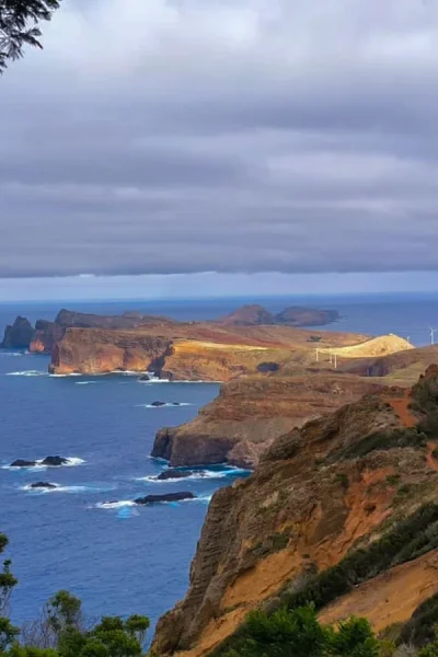 Panoramic coastal view of Madeira’s Ponta de São Lourenço, with rugged red and brown cliffs, scattered sea stacks, wind turbines, and deep blue Atlantic Ocean framed by green foreground trees under a cloudy sky. A Jeep tour experience by Sea La Vie Mountain Expedition.