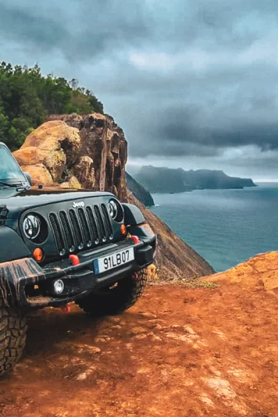 Black Jeep Wrangler parked on a muddy cliff-edge track in Madeira, with dramatic coastal cliffs, forested hills and a moody cloudy sky over the Atlantic Ocean.A Madeira East Mountain Jeep Tour by Sea La Vie Mountain Expedition.