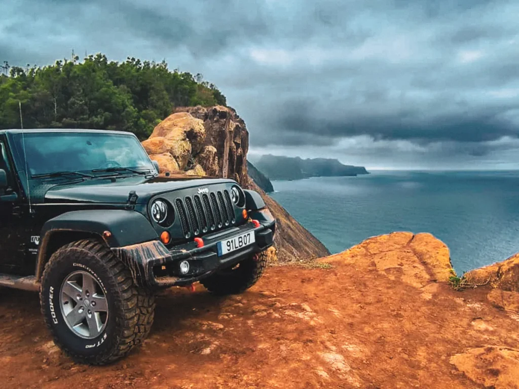 Black Jeep Wrangler parked on a muddy cliff-edge track in Madeira, with dramatic coastal cliffs, forested hills and a moody cloudy sky over the Atlantic Ocean.A Madeira East Mountain Jeep Tour by Sea La Vie Mountain Expedition.