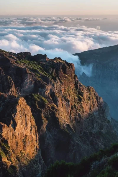Dramatic sunrise view of rugged cliffs in eastern Madeira, with golden light on the rocky slopes and a thick sea of clouds filling the deep valley below. A Jeep tour experience by Sea La Vie Mountain Expedition.