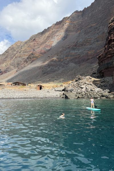 A rugged volcanic shoreline on Desertas Islands with simple wooden huts amid barren slopes, viewed from the sea with a girl on a paddle board and another swimming nearby, an experience by Luxury Charter Madeira.