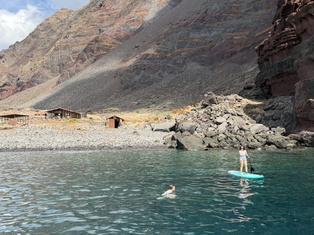 A rugged volcanic shoreline on Desertas Islands with simple wooden huts amid barren slopes, viewed from the sea with a girl on a paddle board and another swimming nearby, an experience by Luxury Charter Madeira.