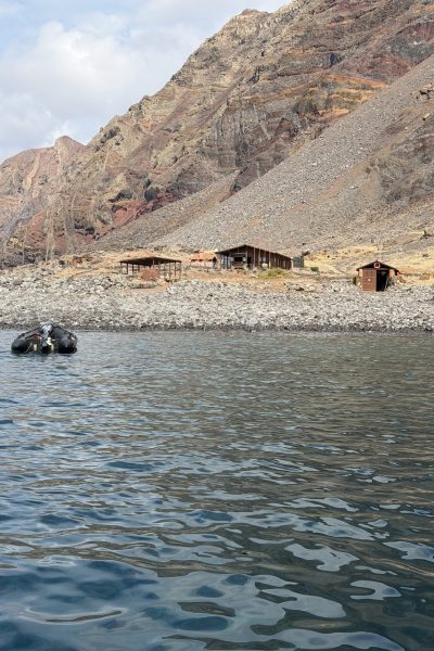 A rugged volcanic shoreline on Desertas Islands with simple wooden huts amid barren slopes, viewed from the sea with two small black buoys floating nearby, an experience by Luxury Charter Madeira.