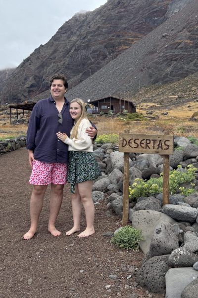 A couple barefoot on a stony path embraces in front of a wooden hut and "DESERTAS" sign, with barren volcanic mountains rising under cloudy skies on Desertas Islands, an experience by Luxury Charter Madeira.