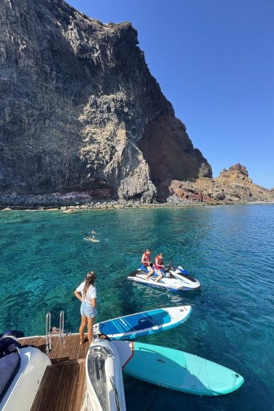 A woman stands on a yacht deck holding a paddleboard, overlooking a blue jet ski and swimmers in turquoise waters beneath massive volcanic cliffs. , an experience by Luxury Charter Madeira.