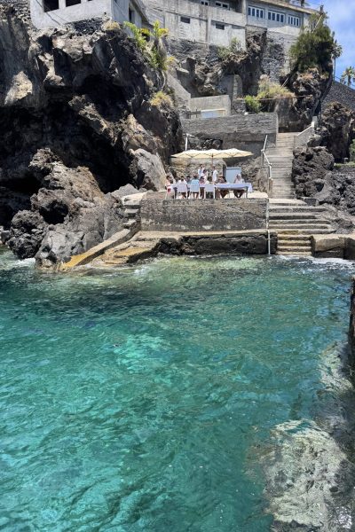 A group dines under white umbrellas at a cliffside restaurant terrace with steps leading to a crystal-clear turquoise sea below, an experience by Luxury Charter Madeira.