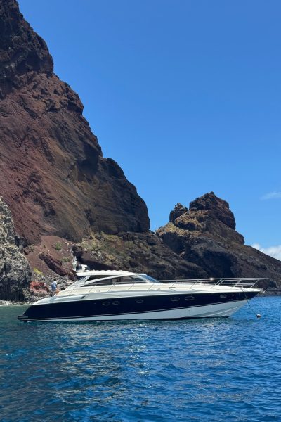 A sleek white and navy yacht floats on clear blue water at the base of towering, rugged volcanic cliffs under a bright, cloudless sky, an experience in a Luxury Yacht Charter near Desertas Island